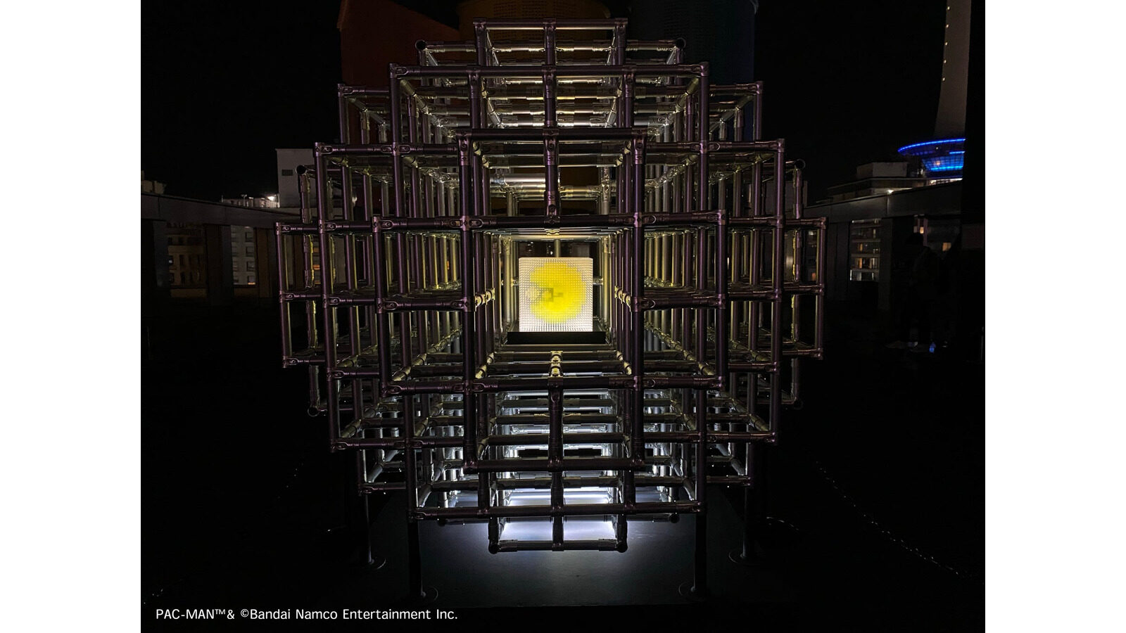 暗い背景に四角い金属フレームが重なり、中央で黄色いパックマンの形が光る立体作品