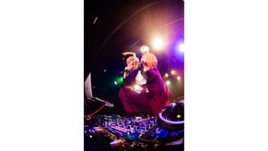 DJ機材の前でマイクを持ちポーズをとるレイチェル